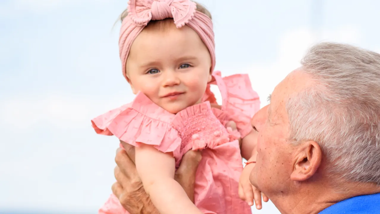 An elderly man holding a baby girl dressed in pink with a matching headband, smiling at her affectionately against a soft blue sky background.