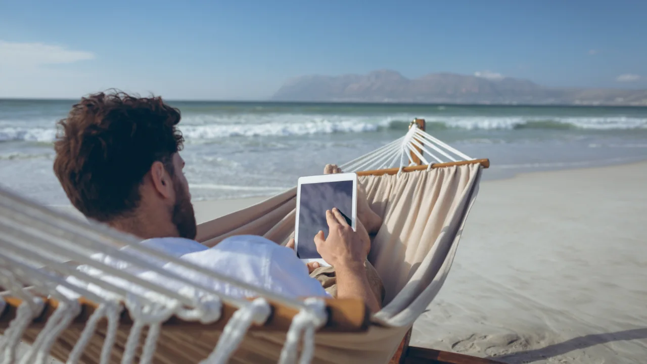 A man relaxing in a hammock on the beach, using a tablet while looking at the ocean, with waves and mountains in the background.