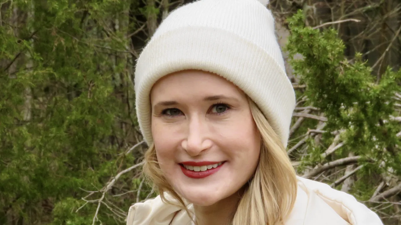 Aimee Bothwell, wearing a white knit hat and light-colored jacket, smiling outdoors with a background of green foliage.