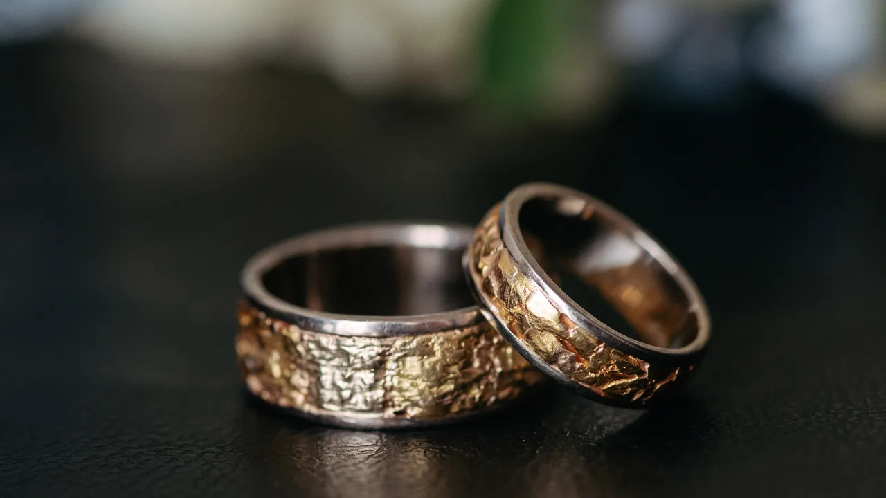 Two textured metal rings with a rustic design, featuring a combination of gold and silver tones, placed on a dark surface.