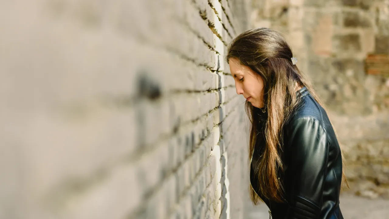 A woman with long hair, dressed in a black leather jacket, standing with her head down against a brick wall, appearing thoughtful or distressed.