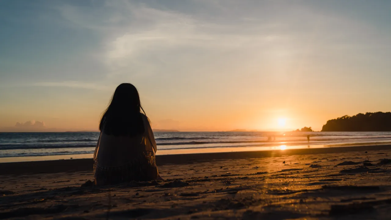 A person sitting on the beach at sunset, looking out at the ocean with the sun setting on the horizon, creating a peaceful and contemplative scene.