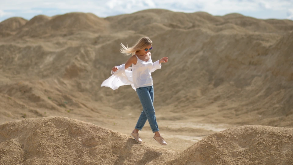 A woman in a white blouse, jeans, and sunglasses playfully balancing on sandy terrain, with barren hills in the background under a partly cloudy sky.