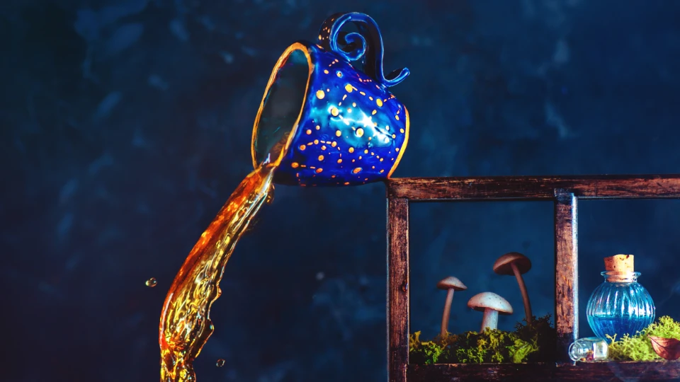 A blue ceramic jug with yellow dots pouring a golden liquid, next to a wooden shelf with moss, small mushrooms, and a blue glass bottle with a cork stopper, set against a dark, moody background.